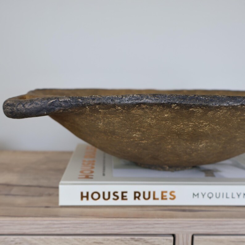 May include: A textured, gold-colored bowl with a dark rim and a handle. The bowl is resting on a book titled "HOUSE RULES" with the author's name, Myquillyn, visible. The bowl is a decorative piece.