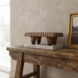 May include: A rectangular, brown, textured wooden riser with four short, thick legs, resting on a white book titled HOUSE RULES by MYQUILLYN SMITH. The riser features a carved, scalloped edge along its top perimeter and a weathered finish.