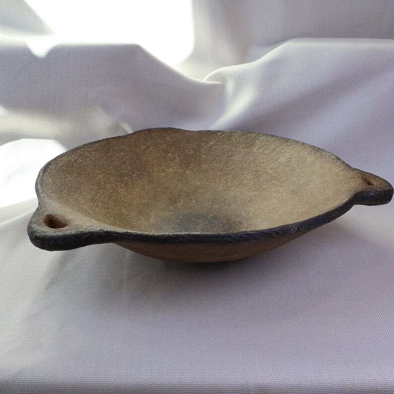 May include: A handmade, oval-shaped bowl with a textured, light brown interior and a dark rim. The bowl has two small, rounded handles on either side. The overall aesthetic is rustic and earthy.