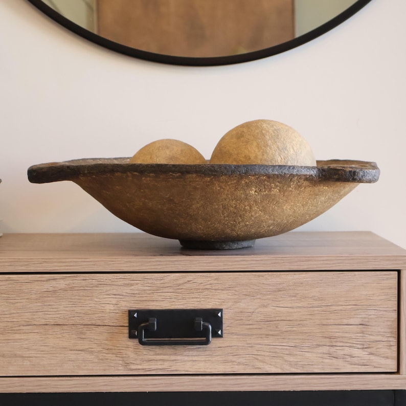 May include: A decorative, handcrafted bowl with a textured, golden-brown finish. The bowl has a wide, shallow design with a dark rim and contains two spherical, light-brown decorative orbs. The bowl is displayed on a wooden surface.