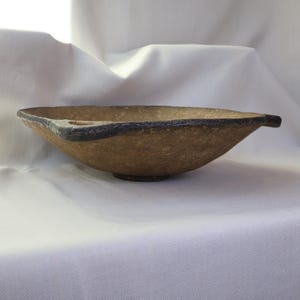 May include: A handmade, textured bowl with a unique shape. The bowl is a light brown color with a dark brown rim and a small, round base. The bowl has a handle on each side. The bowl is likely used for serving food or as a decorative piece.