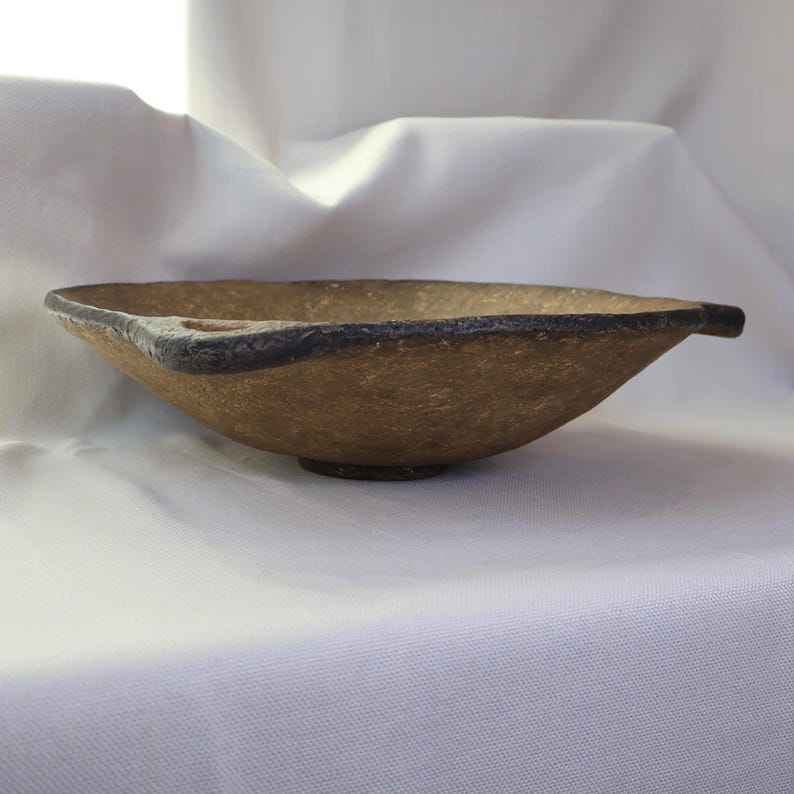 May include: A handmade, textured bowl with a unique shape. The bowl is a light brown color with a dark brown rim and a small, round base. The bowl has a handle on each side. The bowl is likely used for serving food or as a decorative piece.
