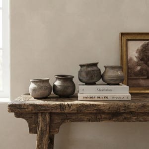 May include: Four small, round, dark-toned ceramic pots with textured surfaces arranged on a rustic wooden table. Two pots sit directly on the wood, while two others rest on a stack of two books titled Minimalista and HOUSE RULES MYQUILLY.
