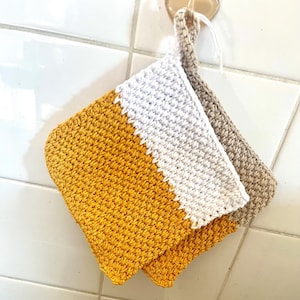 May include: Two handmade crochet potholders. One is mustard yellow with a white diagonal stripe, the other is beige. Both have a loop for hanging. The potholders are hanging on a white tiled wall.