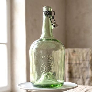 Könnte beinhalten: Eine grüne Glasflasche mit einem Verschluss aus Metall und Keramik. Die Flasche hat einen abgerundeten Körper und einen langen Hals. Sie steht auf einem weißen Sockel. Die Flasche hat ein geprägtes Design auf der Vorderseite.