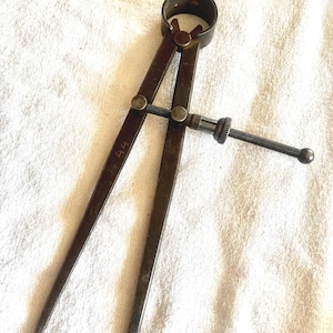 Puede incluir: Un compás de metal vintage con puntas y un ajuste de tornillo. El compás tiene una parte superior circular y está marcado con el número "44". La herramienta está diseñada para medir o transferir distancias.