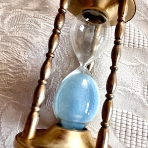 Puede incluir: Un reloj de arena antiguo con marco de latón y arena azul. La bombilla de vidrio está sostenida por cuatro pilares de latón ornamentados. El reloj de arena es un dispositivo clásico para medir el tiempo.