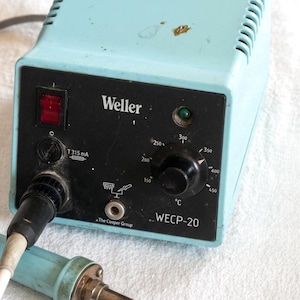 Può includere: Stazione di saldatura Weller azzurra con pannello di controllo nero. Il pannello presenta un interruttore di alimentazione, un quadrante della temperatura e il testo "Weller" e "WECP-20". Un saldatore è collegato.