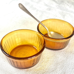 Op de afbeelding: Twee amberkleurige glazen ramekins met een zilveren lepel. De ramekins zijn rond met een geribbeld ontwerp. De lepel is dun met een afgeronde kom. De ramekins staan op een wit oppervlak.