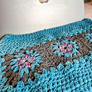 Peut inclure: Une couverture en crochet avec un motif bleu et gris. La couverture est faite de carrés avec un motif de fleur au centre de chaque carré.