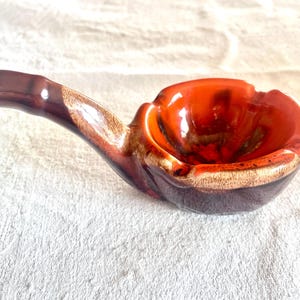 Peut inclure: Un cendrier en céramique en forme de pipe, avec un réceptacle orange foncé et un manche marron foncé. Le cendrier a une finition brillante et un aspect artisanal unique. Le bord du réceptacle est de couleur marron clair.