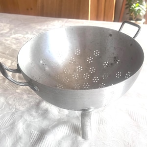 May include: A silver-coloured, vintage aluminium colander with a bowl shape and numerous small drainage holes. It has two curved handles and a single leg for support. The colander shows signs of wear and tear.