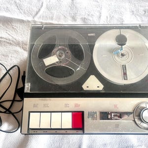 Könnte beinhalten: Vintage Tonbandgerät mit transparentem Kunststoffdeckel. Die Maschine hat silberne und schwarze Akzente mit weißen und roten Knöpfen. Die Tonbandspulen sind unter der Abdeckung sichtbar, und ein Netzkabel ist angeschlossen.