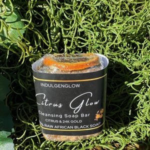 May include: A black and gold bar of soap with the text "Citrus Glow Cleansing Soap Bar Citrus & 24K Gold 100% Raw African Black Soap" on the label.