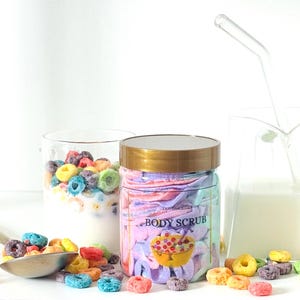 Puede incluir: Un tarro transparente con tapa dorada con la etiqueta "BODY SCRUB" junto a un vaso de cereales y una jarra de leche con pajita. Cereales de colores esparcidos alrededor de una cuchara. El exfoliante corporal tiene un aspecto colorido y en capas.