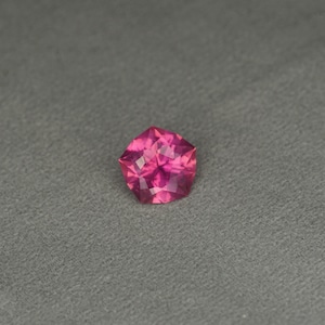 May include: A faceted, pentagon-shaped gemstone in a vibrant shade of pink. The gem is set against a gray background, showcasing its multiple reflective surfaces. The gemstone's cut allows light to reflect, creating a sparkling effect.