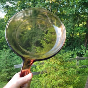 May include: A round, transparent amber-colored glass skillet with a smooth surface and a sturdy handle. The cookware features a circular base and slightly curved walls, showcasing a warm, translucent brown hue that allows light to pass through.