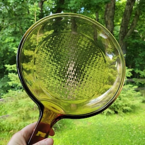 May include: A round, transparent olive green glass skillet with a textured diamond pattern on the interior base. The cookware features a smooth, curved rim and a solid, integrated handle extending from the side, crafted from the same glass material.