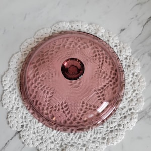May include: A vintage, clear, pink glass lid with a round knob. The lid is resting on a white, crocheted doily with a scalloped edge. The glass lid is a soft, translucent pink color.