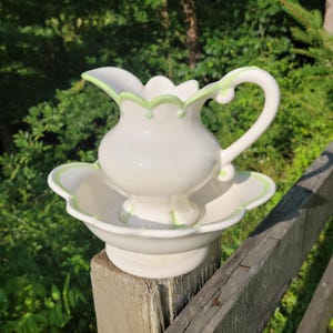 May include: A white ceramic pitcher and matching basin set featuring a scalloped rim design. Both pieces are accented with a thin, light green painted trim along the edges and base, providing a clean, coordinated look for this two-piece set.