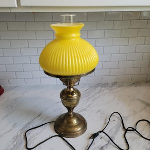 May include: A table lamp featuring a bright yellow, ribbed glass shade with a clear glass chimney top. The base is constructed from aged brass with a circular pedestal and a central stem. A black electrical cord with a plug extends from the base.