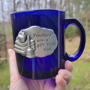 May include: A cobalt blue ceramic mug with a pewter charm attached to the side. The charm features a stack of books, an apple, a bell, and the text "Teachers are a gift from God."