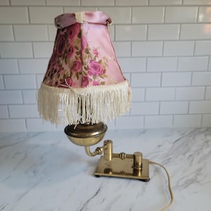 May include: A brass desk lamp with an adjustable arm and a rectangular base. The lamp features a pink fabric shade decorated with a floral rose pattern and finished with a long, cream-colored fringe trim hanging from the bottom edge.