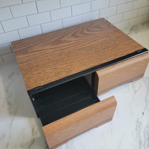 May include: A rectangular wooden desktop organizer featuring a medium brown wood grain finish and a black metal frame. The unit includes two drawers with matching wood fronts, one of which is pulled open to reveal a black interior storage compartment.