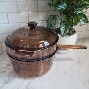 May include: A translucent amber-colored glass saucepan with a matching lid and a long handle. The pot features a fluted, vertical ribbed design along the exterior base and a small, rounded knob on the center of the lid for easy lifting and handling.
