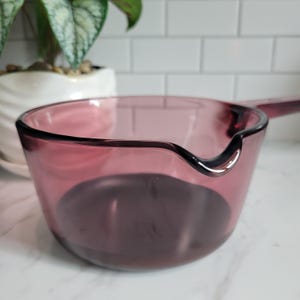 May include: A translucent plum-colored glass saucepan featuring a smooth, rounded bowl, a single integrated handle extending from the side, and a small, curved pouring spout located on the rim for controlled liquid transfer during use.