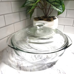 May include: A clear glass casserole dish with a matching lid. The lid has a round, ribbed design and a small handle. The dish is sitting on a white countertop with a plant in the background.