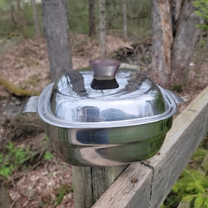 May include: A stainless steel square casserole dish with a lid. The lid has a brown and silver handle. The dish has two side handles. The dish is sitting on a wooden fence post with a natural background.
