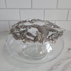 May include: A clear glass bowl with a decorative silver-colored rim featuring a hummingbird and floral design. The bowl is round and sits on a white surface. The rim is detailed with leaves and flowers.