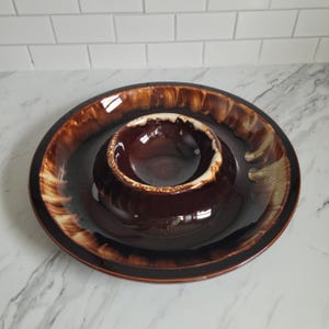 May include: A brown ceramic chip and dip serving set. The set features a large, round outer bowl with a smaller, circular bowl in the center. The bowls have a glossy finish with a brown and tan color scheme.