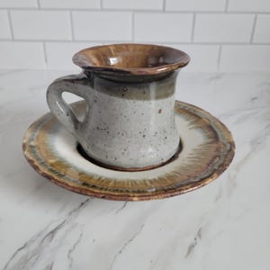 May include: A handmade ceramic mug and saucer set. The mug has a speckled gray body with a brown rim and handle. The saucer has a matching color scheme with a white center and brown and gold edges. The set is designed for serving hot beverages.