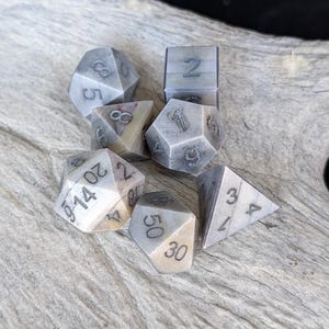 Può includere: Un set di sette dadi poliedrici caratterizzati da una finitura marmorizzata liscia grigio chiaro e bianco sporco con numeri incisi in grigio scuro. La collezione include un d20, d12, d10, d8, d6, d4 e un dado percentuale con cifre ben visibili.