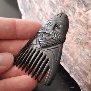 May include: Black wooden comb with a carved face of a man with a beard and a hat. The comb has 7 teeth.