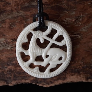 Op de afbeelding: Witte beenderen hanger met een gesneden ontwerp van een mythisch wezen. De hanger is bevestigd aan een zwart koord.