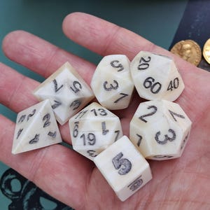 May include: A set of ivory-coloured polyhedral dice with black numbering, including a four-sided die, a six-sided die, an eight-sided die, a ten-sided die, a twelve-sided die, and a twenty-sided die. Two gold coins are also visible.