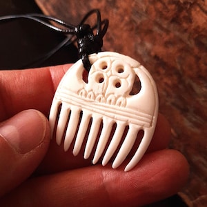 Puede incluir: Collar con colgante de color crema con un diseño de peine tallado. El colgante tiene un patrón detallado con una serie de dientes verticales y una parte superior decorativa con recortes circulares. El collar está ensartado en un cordón negro.