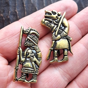 Könnte beinhalten: Zwei goldfarbene Metallbroschen, die einen Krieger und einen Wolf darstellen. Der Krieger hält einen Speer und einen Schild, während der Wolf ein Schwert hält. Beide Figuren tragen Rüstungen und haben detaillierte Gesichtszüge.