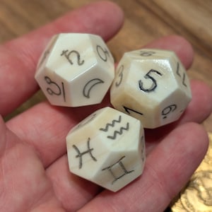 Może przedstawiać: Trzy kości dwunastościenne w kolorze kości słoniowej z czarnymi wytrawionymi symbolami i liczbami. Kości przedstawiają symbole astrologiczne i liczby, trzymane w dłoni. Kości są prawdopodobnie używane do gier fabularnych lub wróżenia.