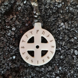 Könnte beinhalten: Weißer runder Anhänger mit einem Kreuzdesign und Runen am Rand. Der Anhänger befindet sich an einer braunen Schnur.