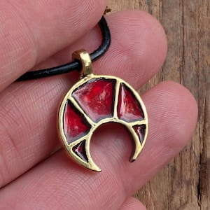 May include: A crescent moon pendant with a gold-colored frame and a red, segmented interior. The pendant is suspended from a black cord. The design is geometric and the overall aesthetic is vintage.