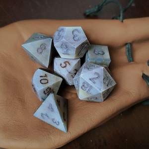 May include: A set of seven polyhedral dice with a marbled white and grey appearance and dark brown numbers, resting inside a tan leather drawstring pouch. The dice include various geometric shapes used for tabletop gaming, featuring clear numerals.