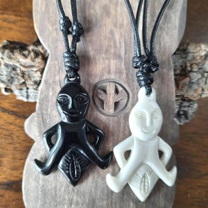 May include: Two pendant necklaces featuring stylized human figures. One is black, the other is off-white. Each figure has a smiling face, outstretched arms, and a leaf-like design at the bottom. The pendants hang from black cords.