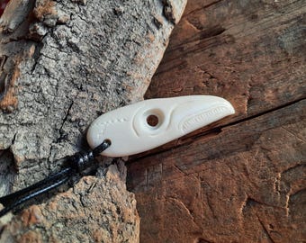 May include: A white carved bone pendant in the shape of a bird's head with a hole for a cord. The pendant is on a black cord and is resting on a piece of wood.