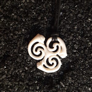 May include: White bone pendant necklace featuring a triple spiral design. The pendant is intricately carved and hangs from a black cord.