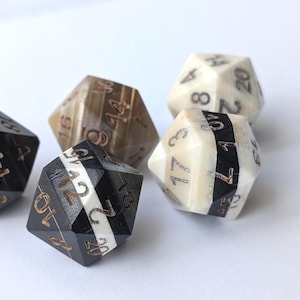 Puede incluir: Cinco dados poliédricos con números. Los dados son de varios colores, incluyendo negro, blanco y marrón. Los números son de un color contrastante, lo que los hace fáciles de leer. Los dados son para juegos de mesa.