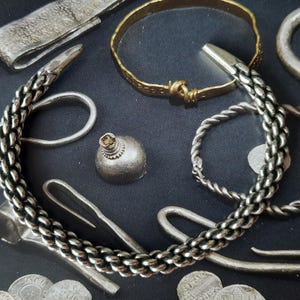 Puede incluir: Un brazalete de plata con un diseño trenzado. El brazalete está sobre una superficie negra con otras piezas de joyería de plata.